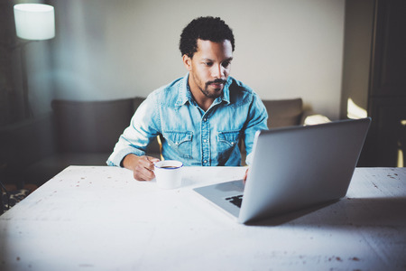 Pensive bearded African man using laptop at home while drinking cup black coffee on the wooden table.Concept of young people work mobile devices.Blurred background.の写真素材