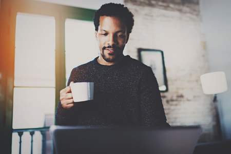 Cheerful African man using computer and smiling at the living room.Black guy holding ceramic cup in hand.Concept of young business people working at home.Blurred background.Selective focus.の写真素材
