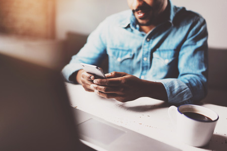 Attractive bearded African man using smartphone while sitting at the wooden table his modern home.Concept of people working with mobile devices.Selective focus on male hands.Blurred background,flares.の写真素材