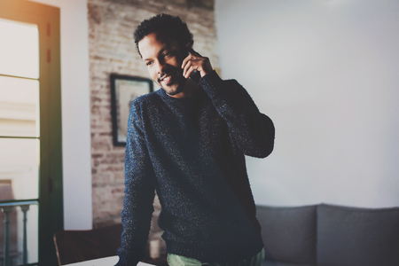 Smiling bearded African man talking on smartphone while standing near the window in his modern apartment.Concept of young business people working at home.Blurred background.の写真素材