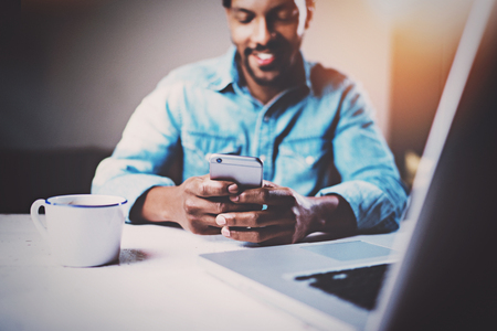 Happy African man using smartphone while sitting at the wooden table his modern home.Concept of young people working with mobile devices.Selective focus on male hands.Blurred background,flares.の写真素材