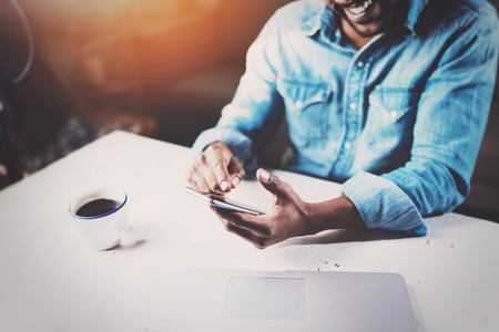 Smiling young African man using smartphone while sitting at the wooden table his modern home.Concept of people working with mobile devices.Selective focus on male hands.Blurred background,flares.の写真素材