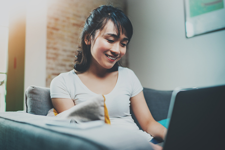 Young Asian woman sitting on sofa at home and using laptop for chatting with friends.Blurred background, flares effect.の写真素材