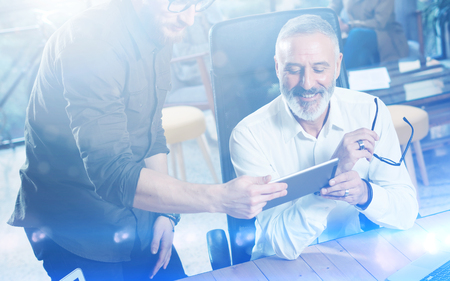 Group of two partners making great idea during work process in modern office.Adult bearded man watching mobile tablet and smiling.Business people meeting concept.Visual effects.の写真素材