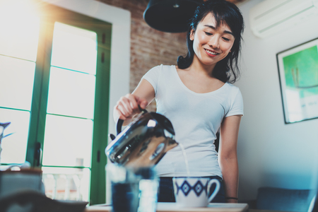 Portrait of young Asian brunette woman using kettle for make tea or black coffee on kitchen at room interior background. Blurred, flares effect.の写真素材