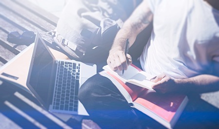 Closeup view of man wearing white tshirt sitting on bench and making notes on text book.Studying at the University,preparation for exams. Books, modern design laptop,backpack.Horizontal,visual effect.の写真素材