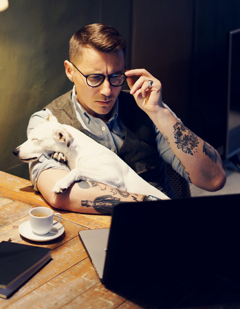 Handsome tattooed man in eyeglasses working at home on laptop while sitting at the wooden table with cute dog on his hands.Adult guy using modern computer for research new business ideas.Vertical.の写真素材