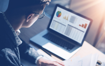 Closeup view of finance market analyst in eyeglasses working at sunny office on laptop while sitting at wooden table.Businessman analyze stock report on notebook screen.Blurred background,horizontal.の写真素材