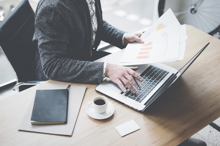 Elegant businessman working at sunny office on laptop while sitting at the wooden table.Man holding documents in his hands.Blurred background.の写真素材