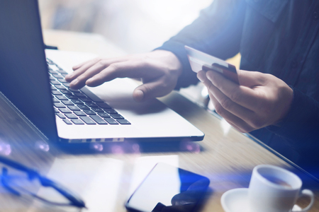 Businessman holding plastic credit card in hand and using laptop computer while sitting at the wooden table.Man making online shopping.Blurred background.Horizontal,flares.の写真素材