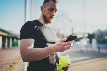 Muscular handsome athlete checking sport results on smartphone application and smart watch after good workout session on city park in the sunny morning.Blurred background.の写真素材