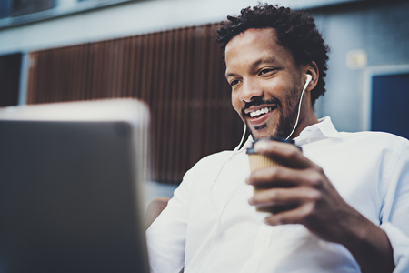 Smiling African American man in headphone making video call via electronic touch pad with take away cup in hand.Concept of guy using Internet-enabled electronic device outdoor.Blurred background.の写真素材
