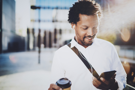 Smiling young African American man in headphone walking at sunny city with take away coffee and using his mobile phone for listen music.Blurred background.の写真素材