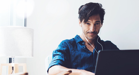 Handsome adult man working at home.Man using contemporary notebook on headphones while sitting in vintage chair.Blurred background. Horizontal wide.Crop.の写真素材