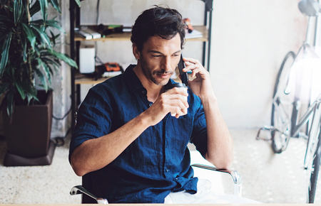 Closeup view of Hispanic elegant businessman working contemporary mobile computer while sitting at the wooden table at sunny office.Man talking on his mobile phone.Blurred background. Horizontalの写真素材