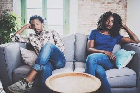 Young displeased black couple.American african men arguing with his stylish girlfriend,who is sitting on sofa on couch next to him with legs crossed.Man looking away offended expression on her face.の写真素材