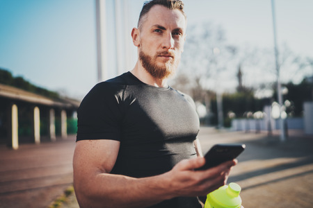 Workout outdoor lifestyle concept.Young man stretching his arm muscles before training.Bearded Muscular athlete using smartphone and exercising outside in sunny park. Blurred background.の写真素材