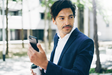 Closeup view of h andsome smiling businessman using smartphone for listining music while walking in city park.Horizontal,blurred background.の写真素材