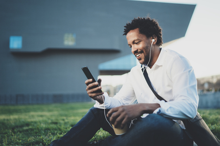 Young African American man in headphone sitting at sunny city park and enjoying to listen to music on his smart phone.Blurred background.の写真素材