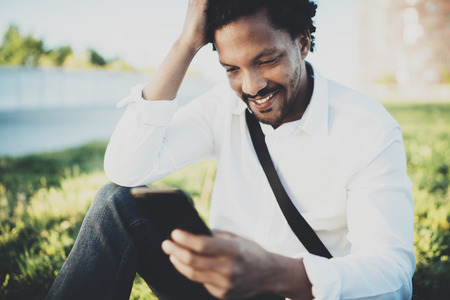 Young bearded African man looking at smartphone in hands while sitting at sunny city park.Concept of happy business people working outside.Blurred background.の写真素材