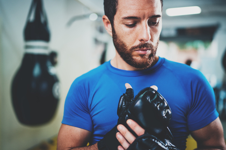 Concentrated Young athlete tying black boxing gloves.Bearded Boxer man prepairing before kickboxing training session in gym. Blurred background. Horizontal.の写真素材