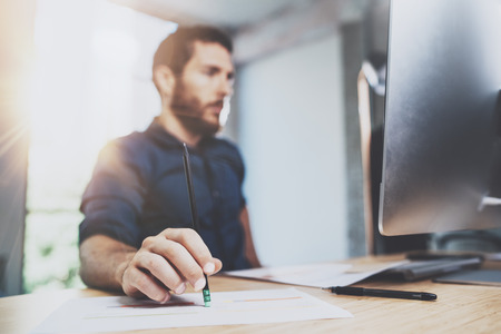 Bearded young coworker working at sunny office.Man using contemporary desktop computer and making notes in notebook.Horizontal.Blurred background.の写真素材