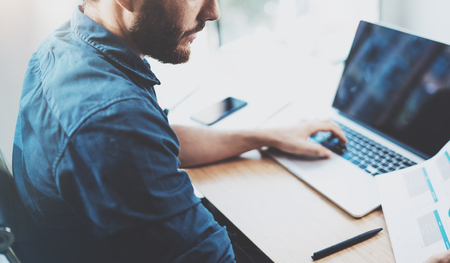 Young pensive man working at sunny office on laptop while sitting at wooden table.Businessman reports on notebook computer.Blurred background,horizontal.Cropped.の写真素材