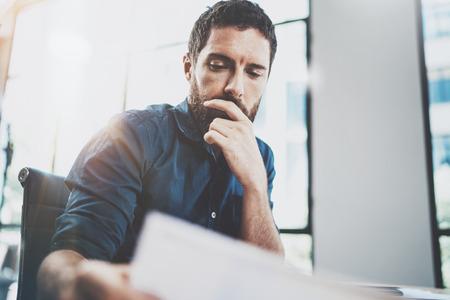 Young bearded man working at sunny loft office on laptop.Businessman holding paper documents hand .Blurred background,horizontal.の写真素材