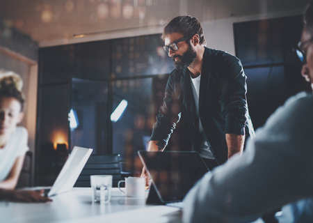 Bearded young coworker working at night office with partners.People using contemporary laptop and modern smartphone.Horizontal.Blurred background.の写真素材
