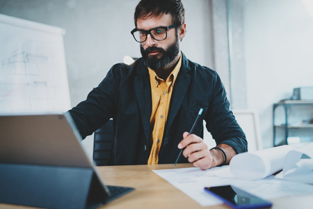 Cropped photo of young bearded male architect wearing eye glasses working on digital tablet dock at his desk. Professional experienced engineer constructionist developer.Horizontal.Blurred background.の写真素材