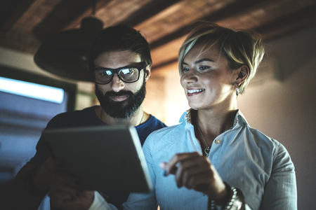 Happy family couple using electronic tablet in livingroom at home.Bearded man in eye glasses making conversation with young blonde woman at night. Blurred background.の写真素材