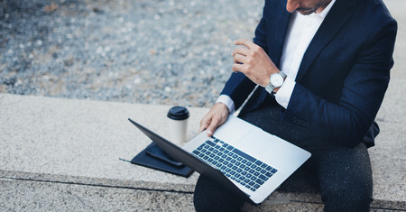 Attractive elegant businessman using contemporary notebook and smartphone sitting outside.Blurred background. Horizontal.Croppedの写真素材