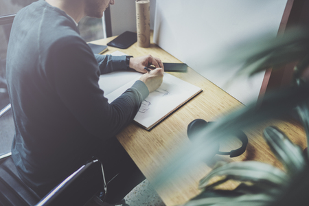 Skilled designer caucasian man drawing abstract sketch with pen.Art work process.Creative hobby.Noting ideas in copy book on workspace desk with papers in modern studio with panoramic windows.Blurredの写真素材