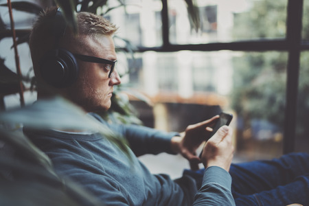 Young man wearing eye glasses and casual clothes listening music in headphones on smartphone at modern place.Horizontal.Blurred background.の写真素材
