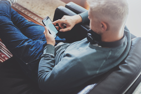 Young man wearing casual clothes sitting on comfort sofa and using smartphone at modern place.Horizontal.Blurred background.の写真素材