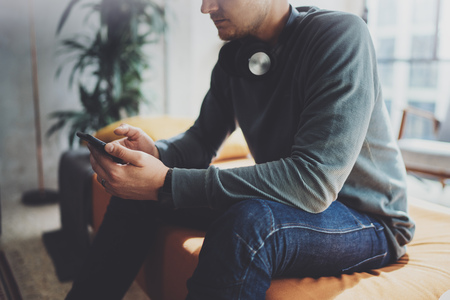 Young hipster wear casual clothes and headphones typing fingers on smartphone screen at modern loft studio.Horizontal.Blurred background.の写真素材