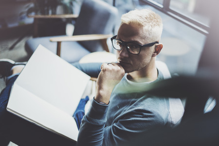 Portrait handsome young man wearing glasses casual clothes holding book hands.Man sitting in vintage armchair modern loft studio reading book. Blurred background.Horizontal.の写真素材