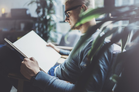Portrait handsome young man wearing glasses casual clothes holding book hands.Man sitting in vintage armchair modern loft studio reading book. Blurred background.Horizontal.の写真素材
