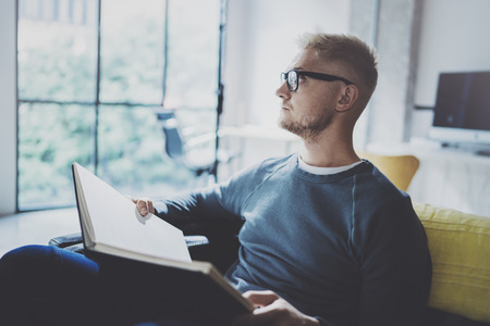 Portrait handsome young man wearing glasses casual clothes holding book hands.Man sitting in vintage armchair modern loft studio reading book. Blurred background.Horizontal.の写真素材
