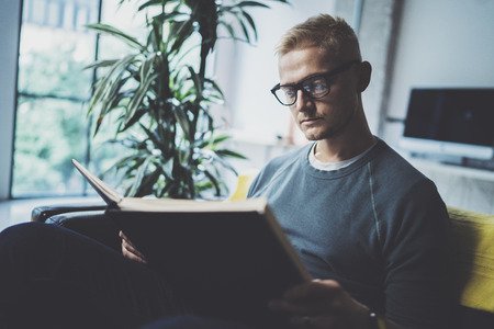 Portrait handsome young man wearing glasses casual clothes holding book hands.Man sitting in vintage armchair modern loft studio reading book. Blurred background.Horizontal.の写真素材