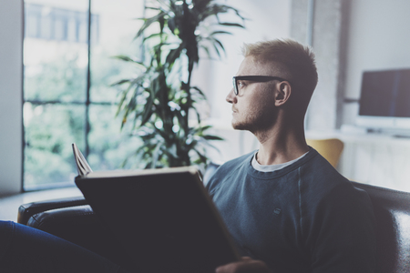 Portrait handsome young man wearing glasses casual clothes holding book hands.Man sitting in vintage armchair modern loft studio reading book. Blurred background.Horizontal.の写真素材