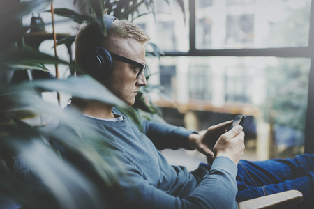 Young hipster wear casual clothes and headphones typing fingers on smartphone screen at modern loft studio.Man using electronic gadgets.Panoramic windows on blurred background.の写真素材