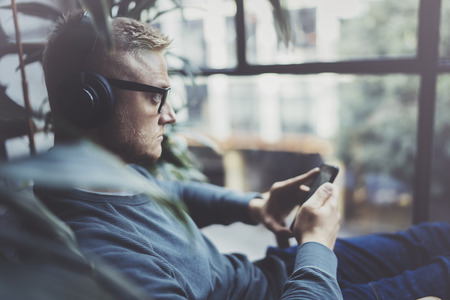 Young hipster wear casual clothes and headphones typing fingers on smartphone screen at modern loft studio.Man using electronic gadgets.Panoramic windows on blurred background.の写真素材