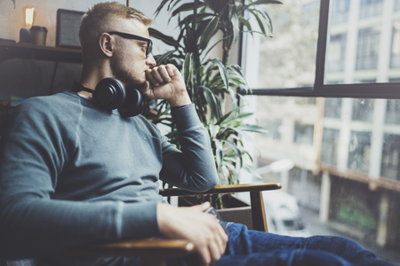 Pensive young man wearing glasses casual clothes.Man sitting in vintage armchair modern loft studio and relaxing whith headphone music.Panoramic windows on blurred background.Horizontal.の写真素材