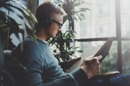 Attractive young man wearing glasses casual clothes.Man sitting in vintage armchair modern loft studio, reading book and relaxing whith headphone music. Blurred background.Horizontal.の写真素材