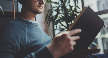 Handsome young hipster wearing glasses casual clothes.Man sitting in vintage armchair modern loft studio, reading book and relaxing whit headphone music. Blurred background.Horizontal.Cropped.の写真素材
