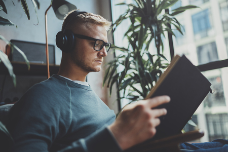 Handsome young man wearing glasses casual clothes.Man sitting in vintage armchair modern loft studio, reading book and relaxing whit headphone music. Blurred background.Horizontal.Cropped.の写真素材