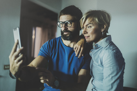 Happy family couple using smartphone in livingroom at home.Bearded man in eye glasses making selfie with young blonde woman at home. Blurred background.の写真素材