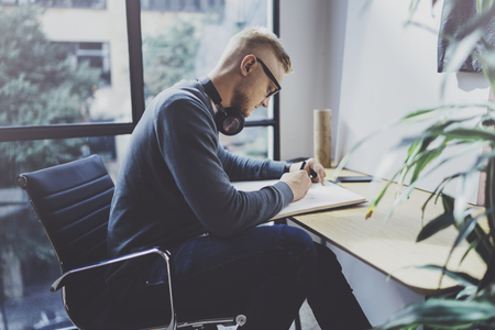Skilled designer caucasian man drawing abstract sketch with pen.Art work process.Creative hobby.Noting ideas in copy book on workspace desk with papers in modern studio with panoramic windows.Blurred.の写真素材