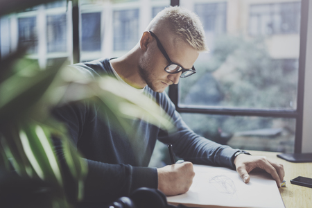 Skilled designer caucasian man drawing abstract sketch with pen.Art work process.Creative hobby.Noting ideas in copy book on workspace desk with papers in modern studio with panoramic windows.Blurred.の写真素材